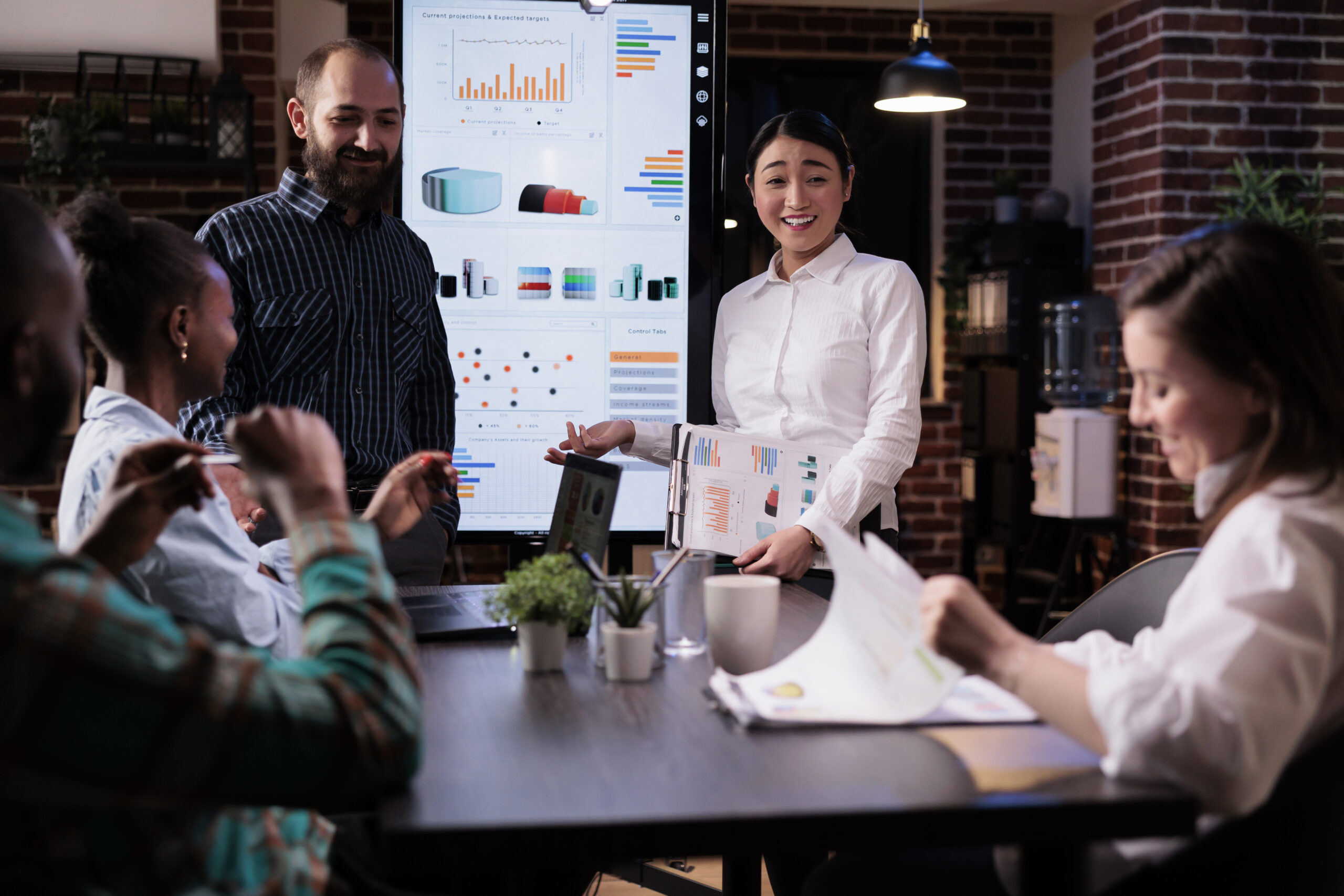 Startup business people talking and smiling at the end of late night sales meeting in corporate office. Marketing team casually discussing marketing strategy in front of wall screen tv with charts.